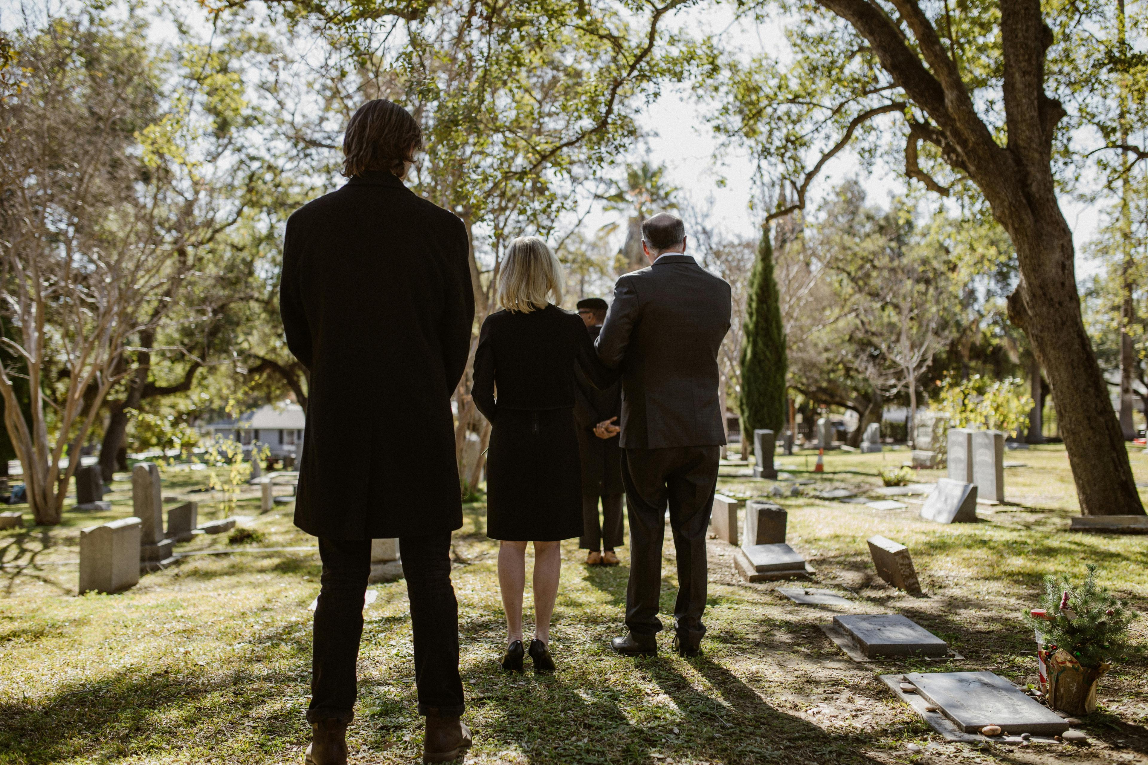 A mourner resting a hand on a flower-laid coffin in a woodland cemetery